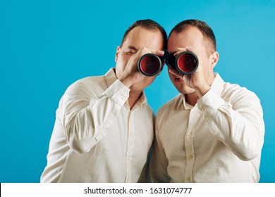 Unrecognizable twin brothers wearing white shirts looking through binoculars at camera, blue wall background
