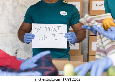 Unrecognizable Volunteer With Medical Face Mask Showing Be Part Of It Sign Board While Other Busy Packing Donations - Concept Of Donate, Help Others During Coronavirus Covid Pandemic