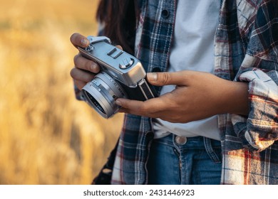 Unrecognizable woman in checkered shirt and casual clothes taking photos with an analog camera in the field at sunset. The image conveys the concept of retro and vintage