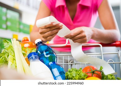 Unrecognizable woman checking a long grocery receipt leaning to a full shopping cart at store. - Powered by Shutterstock