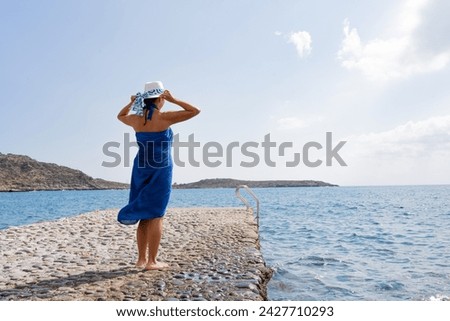 Unrecognizable woman in dark blue summer dress barefoot and holding her hat looking out over the horizon on a beautiful summer morning on a pier on the island of Crete