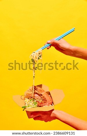 Unrecognizable woman eating delicious hot WOK with shrimps with chopstick from paper box isolated yellow studio background Concept of national food, cuisine, restaurant menu, catering, delivery. Ad