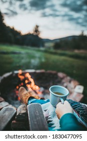 Unrecognizable Woman Enjoying Hot Tea From A Tin Cup In Campsite With Fire Pit. Girl In Folk Blanket By Burning Campfire with mountain landscape with evening sunset sky over the forest and hills.