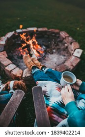 Unrecognizable Woman Enjoying Hot Tea From A Tin Cup In Campsite With Fire Pit. Girl In Folk Blanket By Burning Campfire With Mountain Landscape With Evening Sunset Sky Over The Forest And Hills.