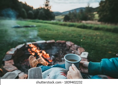Unrecognizable Woman Enjoying Hot Tea From A Tin Cup In Campsite With Fire Pit. Girl In Folk Blanket By Burning Campfire with mountain landscape with evening sunset sky over the forest and hills.