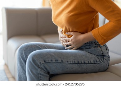 Unrecognizable woman experiencing abdominal pain at home, young female touching her stomach while sitting on couch in living room, showing discomfort and concern, cropped shot, closeup - Powered by Shutterstock