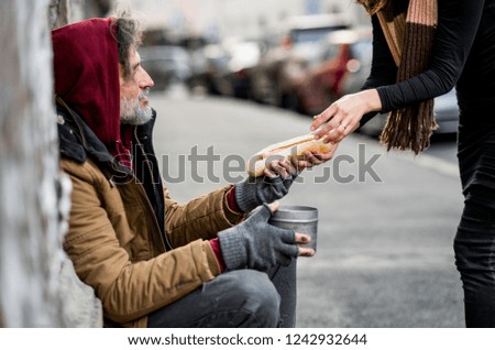 Unrecognizable woman giving food to homeless beggar man sitting in city.