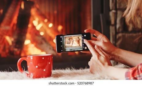 Unrecognizable Woman Hands Taking Cozy Picture on the SmartPhone by the Burning Fireplace - Close Up. Girl making photos of comforting fireside and festive cup scene on a phone