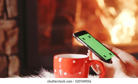 Unrecognizable Woman Hands Using SmartPhone by the Burning Fireplace - Close Up. Female hands with cell phone key-green screen by cozy fireside. Searching internet, using app.