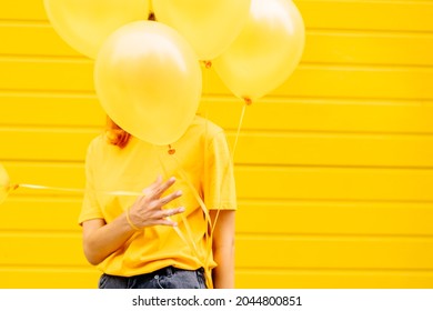 Unrecognizable Woman Hides Her Face Behind Helium Balloons On Yellow Background In City Street Outdoor. Adult Female Holding Ballon In Front Her Face Against Yellow Background, Holiday, Celebration.