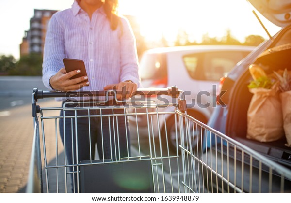 An unrecognizable woman holds up a smart\
phone to view a shopping list as she pushes her cart. Woman with\
shopping cart and shopping list in smartphone\
