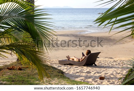 Unrecognizable woman laying down on the wooden longchair at the tropical beach, looking into her phone. 