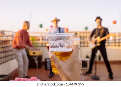 Unrecognizable Woman Lifting And Cheering With A Cold Beer On Intimate Concert. Detail Of A Lager Glass Cheering With Friends. Music And Live Concerts Concept. Intimate Music Group With Summer Vibes.