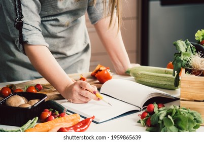 Unrecognizable woman is looking for recipes in cookbook.
				Female chef reading recipes in book and holding pen on table full with fresh vegetables for healthy lunch