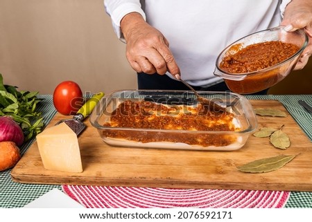 An unrecognizable woman making a lasagna recipe by putting bolognese sauce on top of the preparation with the differents ingredients around the container