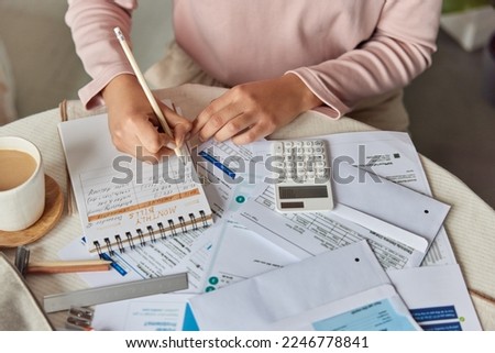 Unrecognizable woman manages household budget or expenditues studies monthly bills does financial paperwork from home sits at table with cup of coffee and calculator writes notes. Reasonable savings