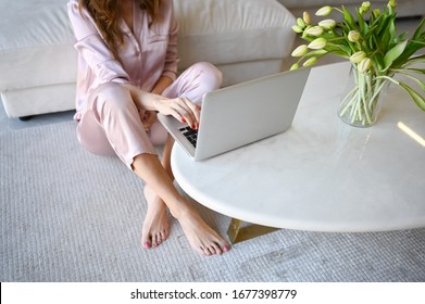 Unrecognizable Woman In Pink Pajamas Sitting On The Floor Working On Laptop. Home Quarantine During Covid-19 Pandemic Corona Virus. Distance Work From Home Concept. White Table With Spring Tulips