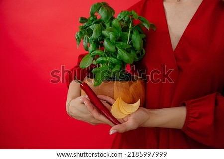 Unrecognizable woman in red clothes holding green plant basil in pot and chili pepper and potato chips in red studio. Choice between fast junk food and healthy eating. Nutrition diet. Copy space