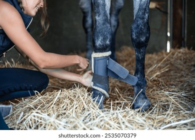 unrecognizable woman rider gently secures leg protectors on her horse inside the stable, showcasing her dedication to the animal's safety and comfort before training - Powered by Shutterstock