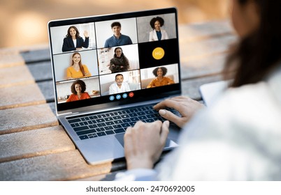 Unrecognizable woman sitting at a desk, typing on a laptop while video conferencing with a diverse group of people displayed on the screen. The individuals on screen are engaged in a virtual meeting - Powered by Shutterstock