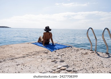 Unrecognizable woman sitting on a towel contemplating the sea from a pier in the south of the Greek island of Crete next to the stairs to go down to bathe