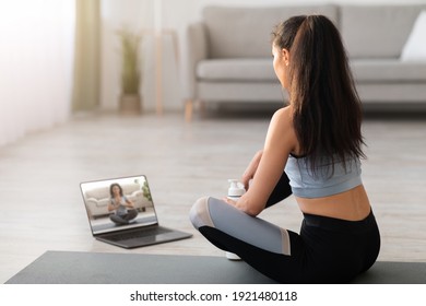 Unrecognizable Woman In Sportswear Sitting On Fitness Mat And Looking At Computer Screen At Home, Having Video Call With Yoga Trainer, Using Laptop. Back View Of Sporty Lady Watching Yoga Class Online