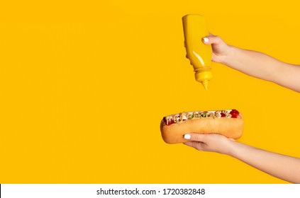 Unrecognizable woman squeezing mustard onto hot dog against orange background, empty space. Panorama