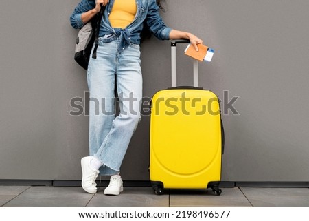 Unrecognizable woman in stylish casual outfit traveller standing over grey background, carrying yellow luggage and backpack, holding passport and flight tickets, cropped, copy space