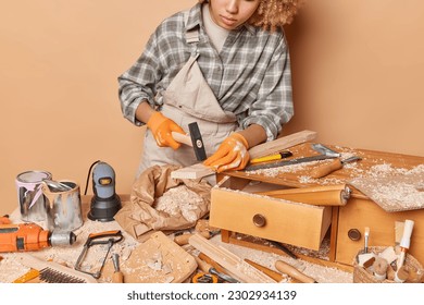 Unrecognizable woman uses hammer uses in workshop creates product made of wood uses tools instruments for woodworking dressed in workwear isolated over brown background. DIY and crafting concept - Powered by Shutterstock