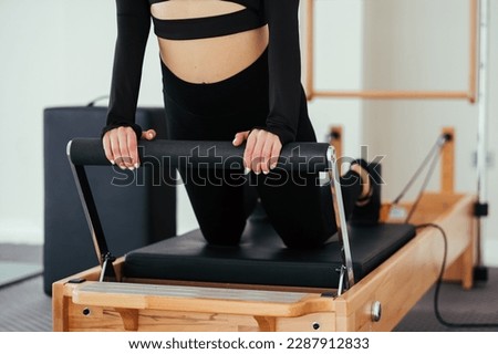 Unrecognizable woman wearing black sportswear practicing pilates exercises on reformer machine in studio.