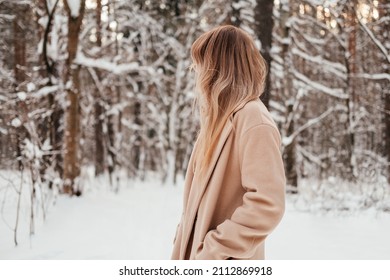 Unrecognizable woman in winter forest looking back. Girl with blonde hair in coat outdoors.