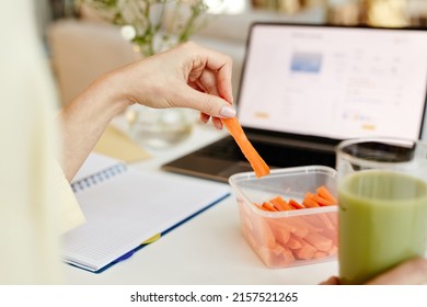 Unrecognizable Woman Working On Laptop In Office And Eating Fresh Sweet Carrot Sticks With Green Smoothie