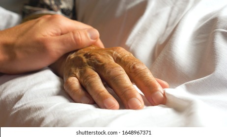 Unrecognizable worried man gently stroking hand of his sick grandmother giving support. Son comforting wrinkled arm of elderly mom lying at bed in hospital. Guy showing care or love to old parent