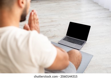 Unrecognizable Young Arab Man Meditating Or Doing Yoga In Front Of Laptop Computer With Blank Screen At Home, Mockup. Millennial Guy Making Namaste Gesture, Greeting Online Personal Trainer On Pc