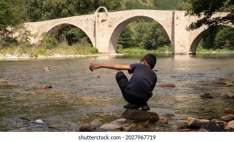 Unrecognizable Young Boy From Behind Playfully Skipping Rocks On The River Water In The Hills Of Southern France. In The Background, A Very Old Stone Bridge Dating From Ancient Rome.