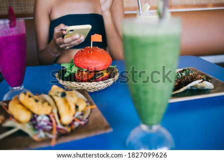 Unrecognizable young female in crop top browsing smartphone while ordering healthy fresh smoothies appetizing hamburger and spicy taco in summer cafe