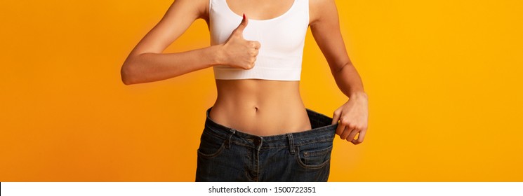 Unrecognizable Young Lady In Oversize Pants Gesturing Thums-Up On Yellow Background. Slimming And Weight Loss. Crop, Panorama, Copy Space