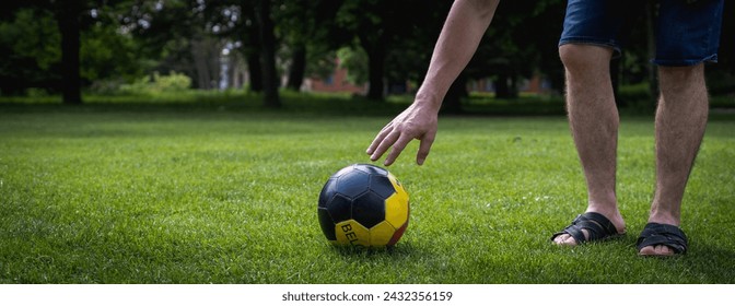 An unrecognizable young man Caucasian man reaches for a soccer ball with Belgium written on it, lying on the green lawn of a city park on a summer day, close-up view from the side. - Powered by Shutterstock