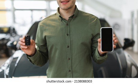 Unrecognizable Young Man Showing New Car Key And Smartphone With Blank Screen At Automobile Dealership Center, Mockup For Auto Rental Mobile App Design. Space For Website