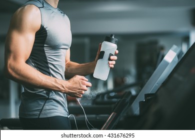 Unrecognizable young man in sportswear running on treadmill at gym and holding bottle of water
