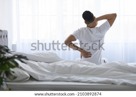 Unrecognizable Young Man Waking Up In Morning With Neck And Back Pain, Millennial Male Having Backache, Massaging Inflamed Aching Zones While Sitting On Bed At Home, Rear View With Copy Space