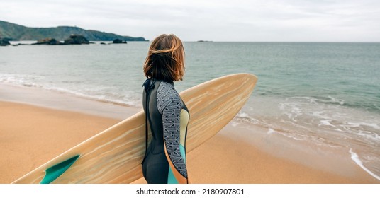 Unrecognizable Young Surfer Woman With Wetsuit Carrying Surfboard Looking To The Sea At The Beach