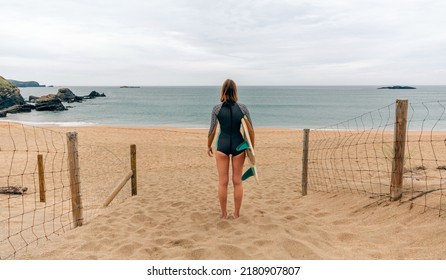 Unrecognizable Young Surfer Woman With Wetsuit And Surfboard Looking At The Sea From The Sand