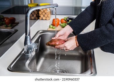 Unrecognizable young woman hands cleaning a sweet potato under water in a home kitchen, in the background there are vegetables. Kitchen concept - Powered by Shutterstock