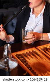 Unrecognizable Young Woman In White Blouse And Suit Drinking Whiskey, Smoking Hookah And Playing Backgammon In Cafe. Business Woman Resting After Work.