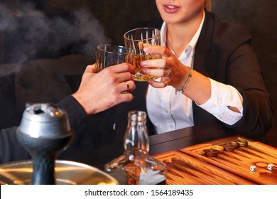 Unrecognizable Young Woman In White Blouse And Suit And Man Drinking Whiskey, Smoking Hookah And Playing Backgammon In Club. Business People Resting After Work.