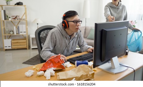 Unrecognized Asian Woman Wife Cleaning House With Garbage Bag Angry For Lazy Partner. Girlfriend Tidy Up Room Messy Dirty Desk Nag Her Boyfriend. Mama Boy Playing Online Video Game On Computer.