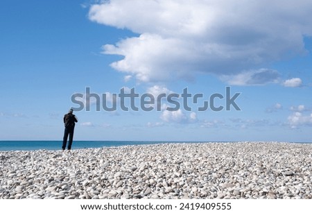 Unrecognized person standing on a pebble beach enjoying the ocean. Cloudy sky, people active outdoors.
