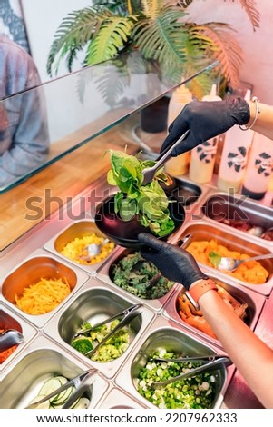 Unrecognized restaurant worker in protective gloves adding vegetables to poke bowl.