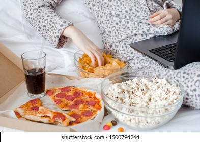 Unrecognized woman eating unhealthy food and crying. Caucasian teenage girl in cute warm pajamas sitting in the bed and watching movie on laptop. Unhealthy overeating lifestyle concept.
